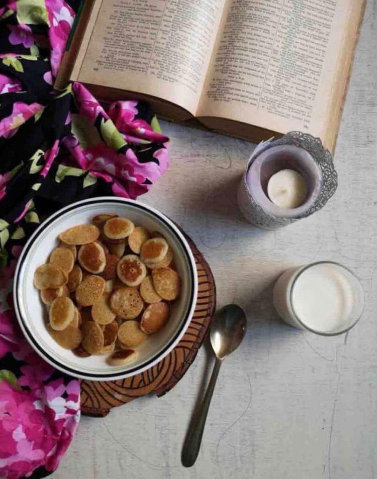 overhead view of Cereal Pancakes served in a white bowl along with a glass of milk , a book and a candle stand in the background.