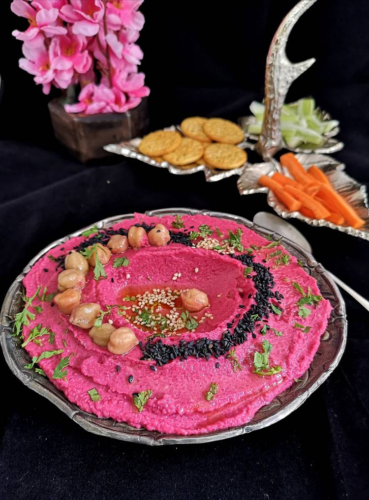 side close up view of Vegan Roasted Beetroot Hummus garnished with black and white sesame seeds, chickpeas, and olive oil, served in a silver bowl, ready to be served with carrot sticks and crackers
