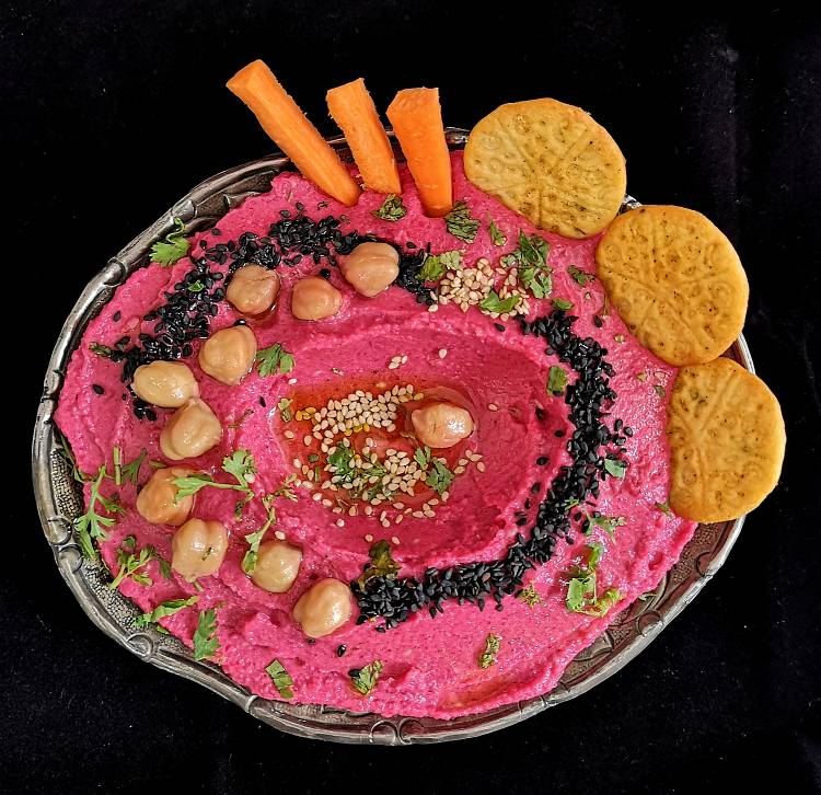 close up view of Vegan Roasted Beetroot Hummus garnished with black and white sesame seeds, chickpeas, and olive oil, served in a silver bowl, ready to be served with carrot sticks and crackers