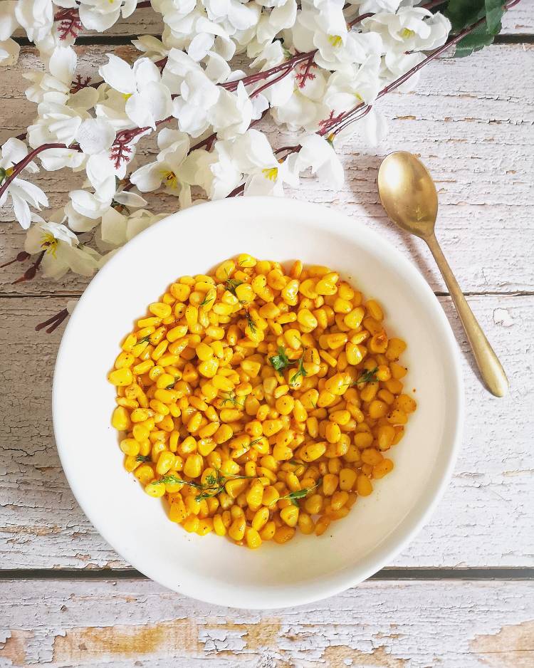 Overhead Photo of buttered corn presented on a wooden white board served in a deep white ceramic bowl with a brass spoon, and dry white flowers