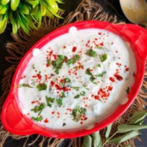 top view of vrat wale cucumber raita recipe garnished with coriander leaves and red chili powder / Recipe of cucumber raita / how to make cucumber raita