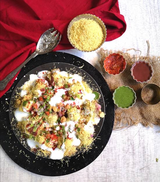 top view image of Dahi bhalla papdi chaat recipe served with garlic chutney, khajur, dates chutney, green coriander chutney, nylon sev with a silver spoon and red cotton napkin.
