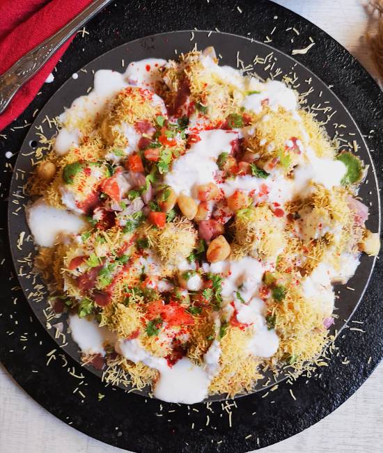 overhead photo of Dahi papdi chaat garnished with chickpeas, pomegranates, coriander leaves, nylon sev and dahi and presented on a black plate